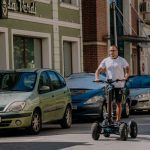 Верига на градска уличка с паркирани автомобили и човек каращ електрически тротинет.